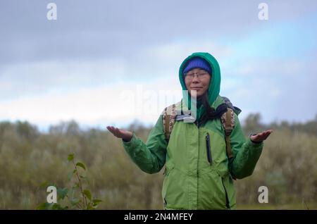 Yakut asiatique fille touriste dans un chapeau de veste et la capuche et avec un sac à dos est méditant, fermant ses yeux, sous la grêle et la pluie levant ses mains à nouveau Banque D'Images