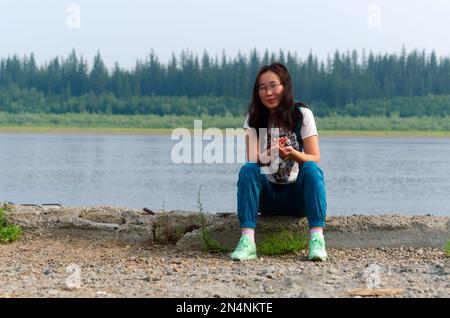 Sports gaies Yakut jeune fille asiatique assis sur la rive de pierre de la rivière Viluy dans la forêt du Nord met ses mains dans le château et sourit. Banque D'Images