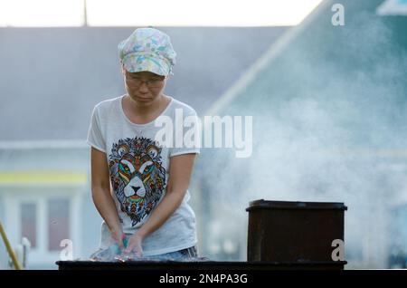 Yakut asiatique fille avec des verres et un chapeau prépare un barbecue à un grand grill dans la fumée sur le fond d'une maison privée résidentielle dans le Nord Banque D'Images