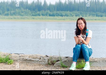 Frustré Yakut fille voyageur asiatique criant avec les mains pliées sur la banque de pierre de la rivière du Nord et la forêt dans le nord de la taïga russe Banque D'Images