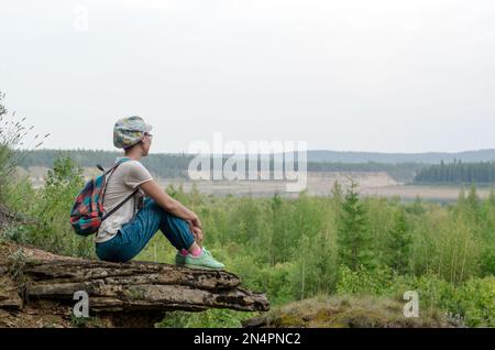 Yakut asiatique fille touriste avec un sac à dos et une casquette assis sur le bord de la montagne dans la toundra sauvage de Yakutia embrassant ses jambes de repos et regarder Banque D'Images