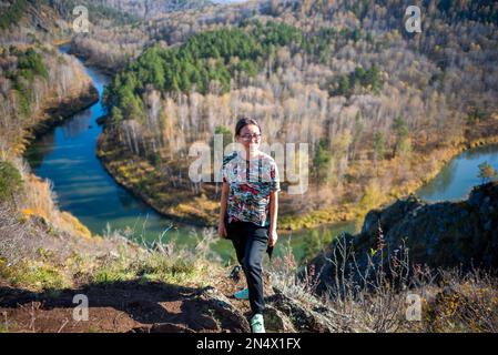 Yakut asiatique fille en lunettes dans un t-shirt et avec un sac à main posant souriant debout sur le côté de la montagne avec un tour de rivière au fond de la Banque D'Images