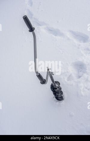 Une perceuse à glace à main noire en fer avec une poignée incurvée et des vis congelées dans de la glace est vissée dans la glace en hiver sur un fond de sn blanc Banque D'Images