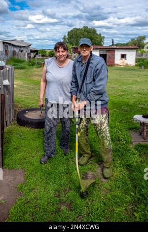 Famille dans le jardin dans le village d'une femme ébourieuse et d'un frère mince avec un support de coupe-herbe sur la pelouse en été. Banque D'Images