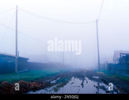 Une grande flaque sur une rue de village le matin brouillard épais entre les maisons avec des clôtures et des poteaux avec des fils électriques dans le nord de Yakutia. Banque D'Images