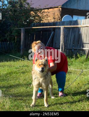 Une joyeuse Yakut grosse femme en lunettes pose avec son chien Akita inu sur une laisse sur l'herbe verte d'un village de Yakutia. Banque D'Images