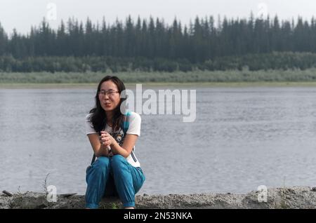 Sports surpris Yakut asiatique avec des lunettes jeune fille assise sur l'ancien quai de la rivière Viluy dans la taïga du nord avec sa main dans le château Banque D'Images