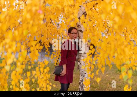 Yakut asiatique jeune fille heureuse robe avec sac à main avec des verres et avec des ornements embrassant le bouleau avec des feuilles jaunes en automne. Banque D'Images