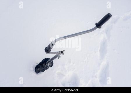 Une perceuse à glace à main noire en fer avec poignée incurvée se visse dans la glace en hiver sur fond de neige blanche. Banque D'Images