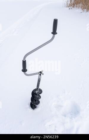 Une perceuse à glace à main noire en fer avec une poignée incurvée et des vis congelées dans de la glace est vissée dans la glace en hiver sur fond blanc Banque D'Images