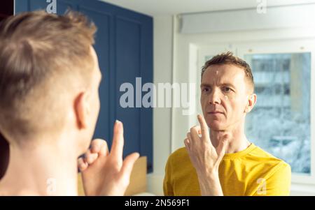 Homme mûr appliquer de la crème hydratante avec le doigt sur les zones claires du visage. Portrait masculin dans le miroir de la chambre. Soin de la peau routine matinale, beauté soignée Banque D'Images