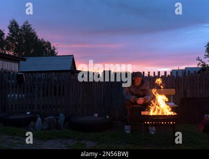 Un vieil homme s'assoit seul sur un banc dans le terrain du village derrière la clôture de la maison en regardant le feu dans le grill le soir au coucher du soleil. Banque D'Images