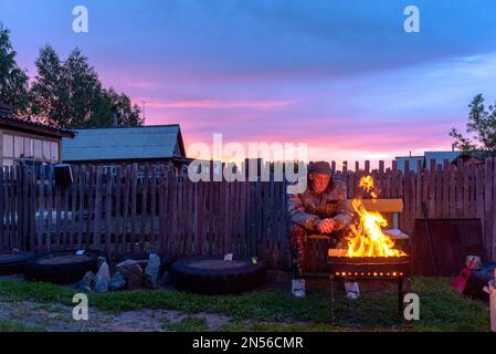 Un vieil homme solitaire s'assoit malheureusement sur un banc dans une maison de village derrière une clôture qui donne sur le feu d'un feu de barbecue au coucher du soleil dans la soirée. Banque D'Images