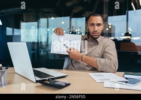 Portrait déçu homme d'affaires à l'intérieur du bureau, homme afro-américain montrant un graphique de document avec de mauvaises performances financières à l'appareil photo, financier en sous-couche travaillant à l'intérieur du bureau avec un ordinateur portable. Banque D'Images