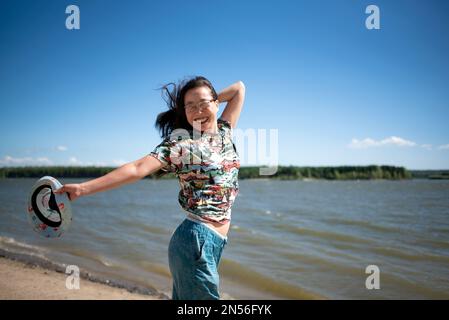 Un joyeux sport Yakut asiatique fille en lunettes et un t-shirt sur la rive d'une rivière venteuse avec des vagues tenant ses cheveux et un chapeau dans son autre main. Banque D'Images