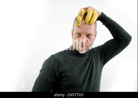 photo comique d'un homme tenant trois bananes gâtées sur sa tête comme une coiffure les yeux squited scald tête rouge avec une barbe grise et la moustache vert chandail blanc fond faire des visages faire des visages Banque D'Images