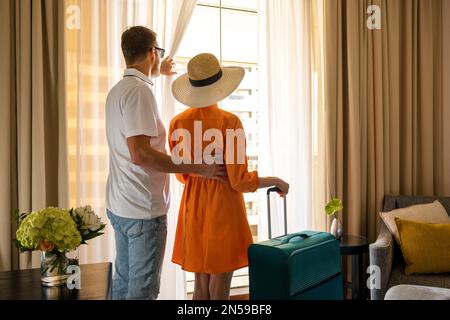 un couple de jeunes voyageurs avec des bagages qui regardent de la fenêtre de la chambre d'hôtel après l'arrivée Banque D'Images