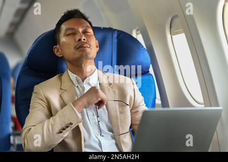 Homme d'affaires millénaire distrait du travail informatique tout en étant assis confortablement assis dans la cabine d'avion Banque D'Images