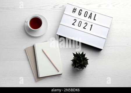 Boîte lumineuse avec hashtag objectif 2021 près des carnets et une tasse de thé sur une table en bois blanc, plat. Objectifs de la nouvelle année Banque D'Images