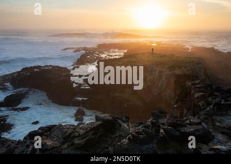 Une personne se tient sur un éperon côtier rocheux alors que de grandes vagues se brisent contre la rive au coucher du soleil, Chatham Island, Nouvelle-Zélande, janvier Banque D'Images