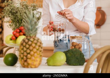 gros plan des mains de l'hôtesse tenant des noix dans un pot et des légumes couchés, des fruits au premier plan sur la table de cuisine. cuisson. Banque D'Images