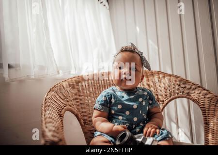 Petite fille en robe bleue à pois a une émotion positive assise sur une chaise de canne et ombre sur le visage. Bébé afro-américain détend un Banque D'Images