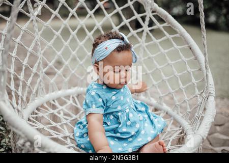 Petite fille en robe bleue à pois a une émotion positive assis sur une corde de cocon en coton macrame blanc suspendue. Bébé afro-américain rel Banque D'Images