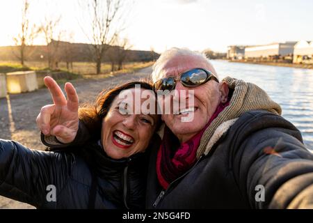 couple d'âge moyen portant des vêtements d'hiver prenant un selfie sur les rives de la rivière - concept de personnes dans les loisirs Banque D'Images