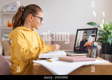 Fille d'école ayant vidéo conférence avec professeur en ligne sur ordinateur portable dans le salon à la maison. Homeschooling, formation à distance, formation en ligne due à une pandémie de coronavirus. Vue latérale. Concept d'apprentissage en ligne Banque D'Images