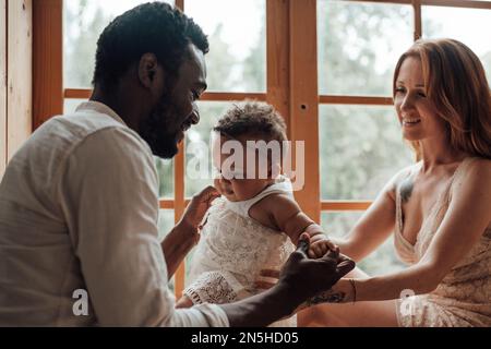 Famille de race mixte avec la peau sombre bébé fille passer du temps ensemble utiliser la fenêtre claire dans l'appartement Snug. Homme afro-américain sa femme de peau équitable an Banque D'Images