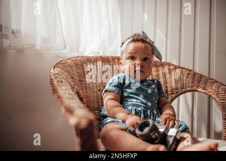 Petite fille en robe bleue à pois a une émotion positive assise sur une chaise de canne et ombre sur le visage. Bébé afro-américain détend un Banque D'Images