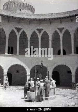 1963, historique, visiteurs du château de Bellver, Palma, Majorque, à l'intérieur de la cour intérieure circulaire, debout au puits d'eau. Construite pour le roi Jacques II de Majorque au XIVe siècle, la forteresse de style gothique est le seul château circulaire existant en Espagne. Banque D'Images