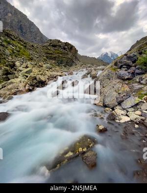 Le ruisseau alpin coule sur des pierres près des montagnes avec des glaciers et de la neige en Altai, en Sibérie, derrière les nuages pluvieux pendant la journée. Cadre vertical. Banque D'Images