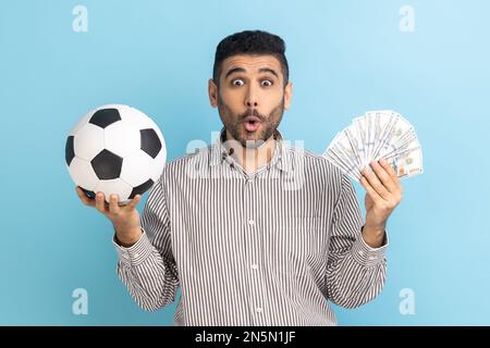 Un homme d'affaires stupéfait tenant le ballon de football et fan de billets de dollars, Paris sportifs, grand gain, regardant la caméra, portant une chemise rayée. Studio d'intérieur isolé sur fond bleu. Banque D'Images