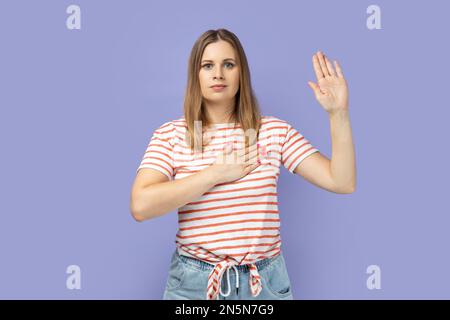 Portrait d'une femme blonde patriotique sérieuse portant un T-shirt rayé levant un bras et mettant sur la poitrine un autre faisant serment, prestation de serment. Studio d'intérieur isolé sur fond violet. Banque D'Images