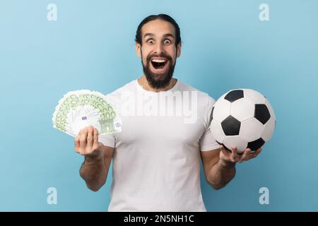 Portrait d'un homme avec barbe portant un T-shirt blanc tenant le ballon de football et des centaines d'euros de factures, criant de regarder l'appareil photo, Paris et gagner. Studio d'intérieur isolé sur fond bleu. Banque D'Images