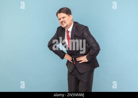 Portrait d'un malade malsain malade malade malade avec moustache ayant une forte douleur dans son estomac, touchant le ventre, frognant le visage, portant un costume noir avec une cravate rouge. Prise de vue en studio isolée sur fond bleu clair Banque D'Images