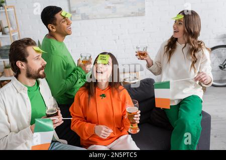 Amis multiethniques avec des notes collantes sur les fronts tenant des boissons alcoolisées et jouant à Guess Who Game le Saint Patrick Day Banque D'Images