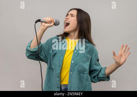 Portrait d'une femme chantant des chansons dans un microphone, performance de chanteur, garde les yeux fermés, portant une veste de style décontracté. Prise de vue en studio isolée sur fond gris. Banque D'Images