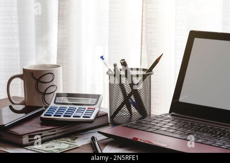 Un bureau en face se une fenêtre où sont un ordinateur portable, une calculatrice, un agenda, des documents, des stylos, argent et une tasse. Banque D'Images