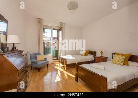 Chambre avec deux lits simples en bois, bureau avec tiroirs et capote fermée, fauteuil bleu et balcon avec vue Banque D'Images