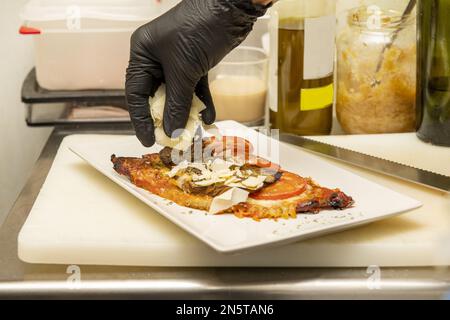 Un chef aux mains gantées de plastique noir mettant les dernières touches de copeaux de parmesan sur un milanais Banque D'Images