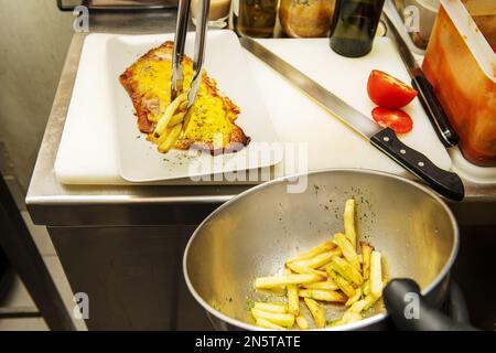 Un chef aux mains gantées de plastique noir mettant la touche finale avec des frites sur une milanesa dans une cuisine de restaurant Banque D'Images