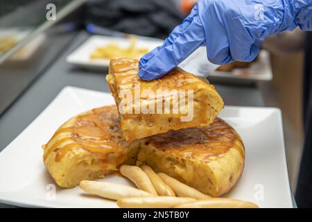 Un chef avec des mains gantées en plastique bleu séparant un morceau de tortilla espagnole pour préparer un tapa Banque D'Images