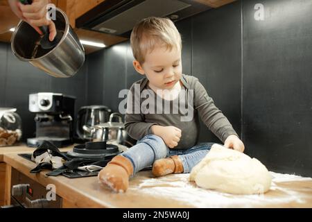 Petit garçon tranquille, agréable, enfant goûtant et touchant le caillot de pâte, assis sur un tiroir en bois dans la cuisine. Pâtisserie artisanale Banque D'Images