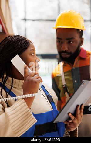 Femme afro-américaine tenant une tablette tout en parlant sur un téléphone fixe dans un entrepôt. Le responsable de la logistique consulte l'employé du magasin et répond à l'appel du superviseur dans la salle de stockage Banque D'Images
