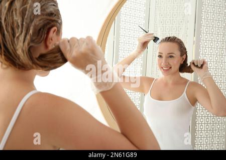 Jeune femme appliquant de la teinture sur les cheveux près du miroir à l'intérieur Banque D'Images