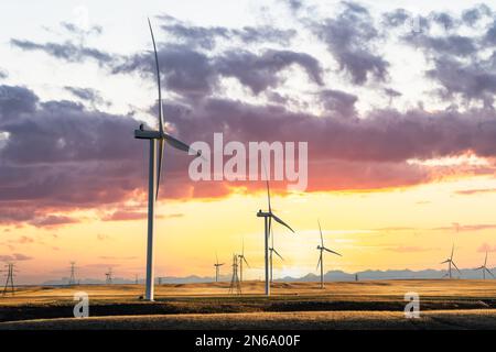 Moulins à vent au coucher du soleil produisant de l'énergie verte surplombant les champs de blé agricole sur un paysage de prairie avec des montagnes éloignées sous un couleurs spectaculaire Banque D'Images