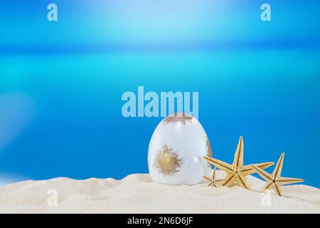 Oeuf d'or blanc avec étoiles de mer sur une plage de sable de la mer ou de l'océan. Carte postale de vacances dans un pays ensoleillé. Joyeuses Pâques. Voyage, printemps, été. Copier l'espace Banque D'Images