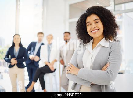 Portrait d'une jeune femme d'affaires confiante de race mixte debout avec ses bras croisés dans un bureau avec ses collègues en arrière-plan. Ambitieux Banque D'Images